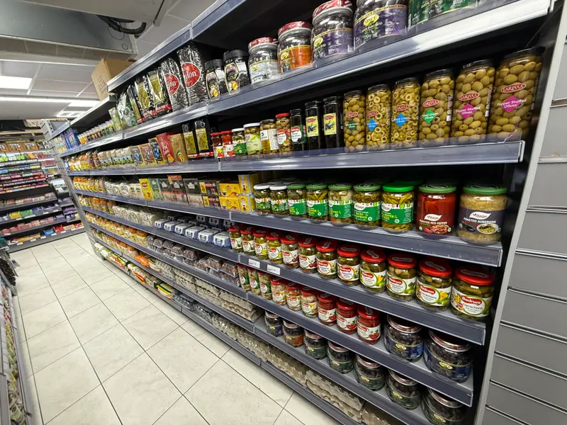 Shelves of jarred olives, pickles, roasted peppers and preserves along a shop aisle