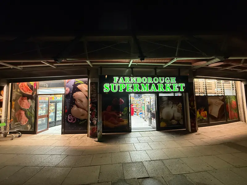 The Farnborough Supermarket shopfront at night, with its green illuminated sign and window displays of fresh meat and groceries